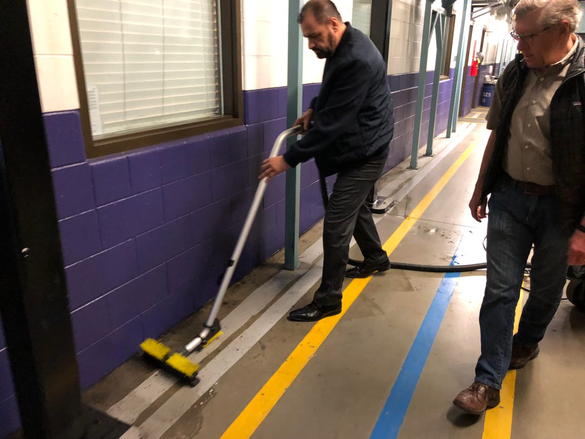 Two men in a service corridor during a floor scrub — one running an edge brush along the base of the block wall, the other walking the wet floor beside painted yellow and blue aisle lines