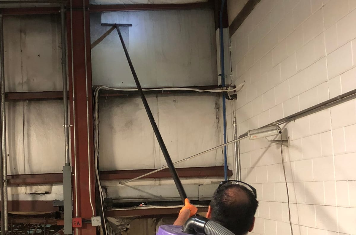 A worker in ear protection running a backpack-vacuum pole up a soot-streaked wall panel beside a white block wall