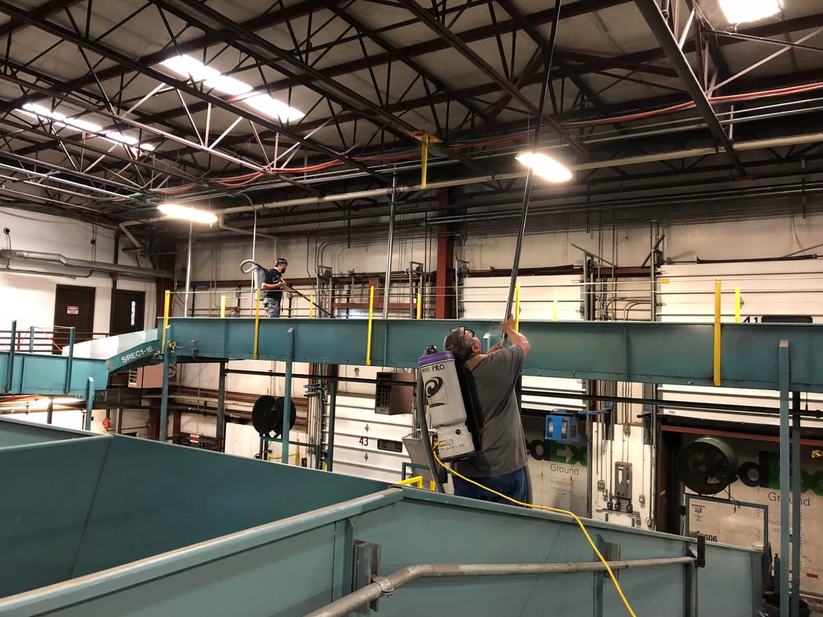 A worker with a backpack vacuum running an extension pole along overhead steel and ductwork above teal plant machinery