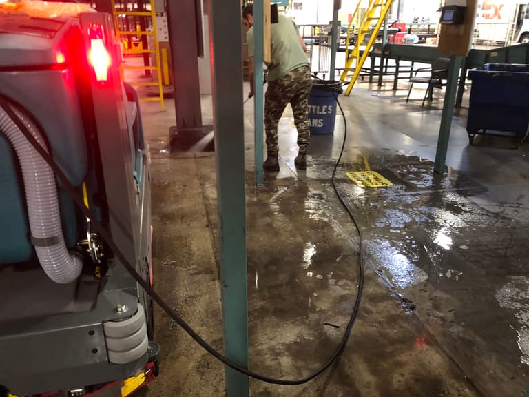 An auto-scrubber parked on a wet warehouse dock floor while a worker washes down between the columns behind it