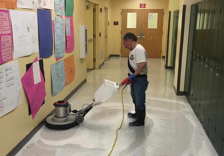 A worker running a floor machine down a school corridor lined with lockers and a bulletin board, the floor wet and stripped back