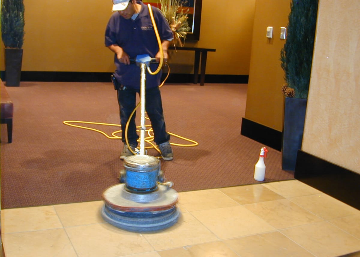 A technician running a burnishing machine across a stone lobby floor beside a yellow wall