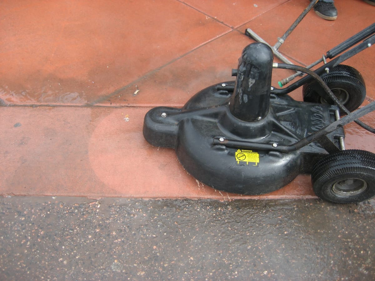 A rotary surface cleaner resting on colored concrete, a hard line dividing the washed slab from the untouched one